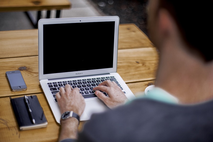 picture of a man sitting on a wooden table in front his computer beside his cellphone and minimal notebook with pen 