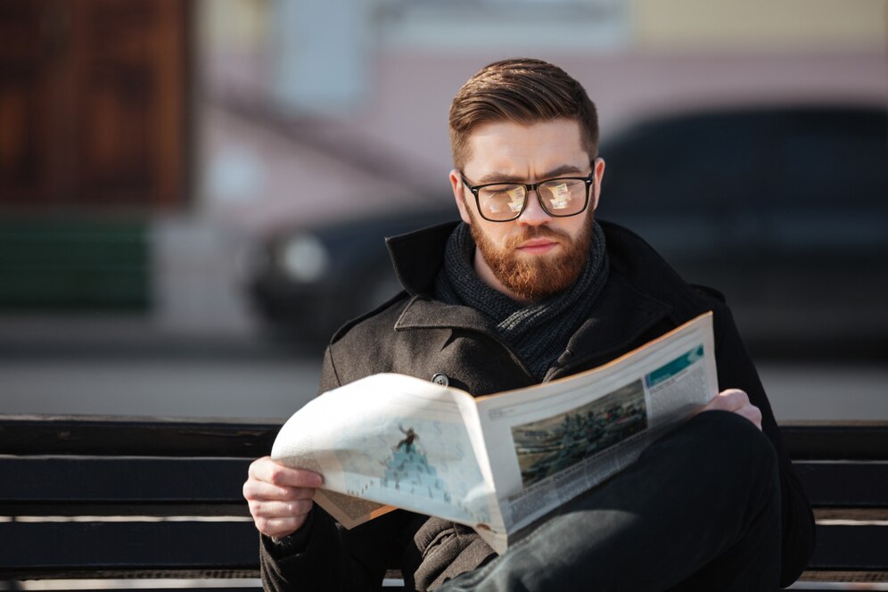 concentrated young man sitting bench reading newspaper outdoors