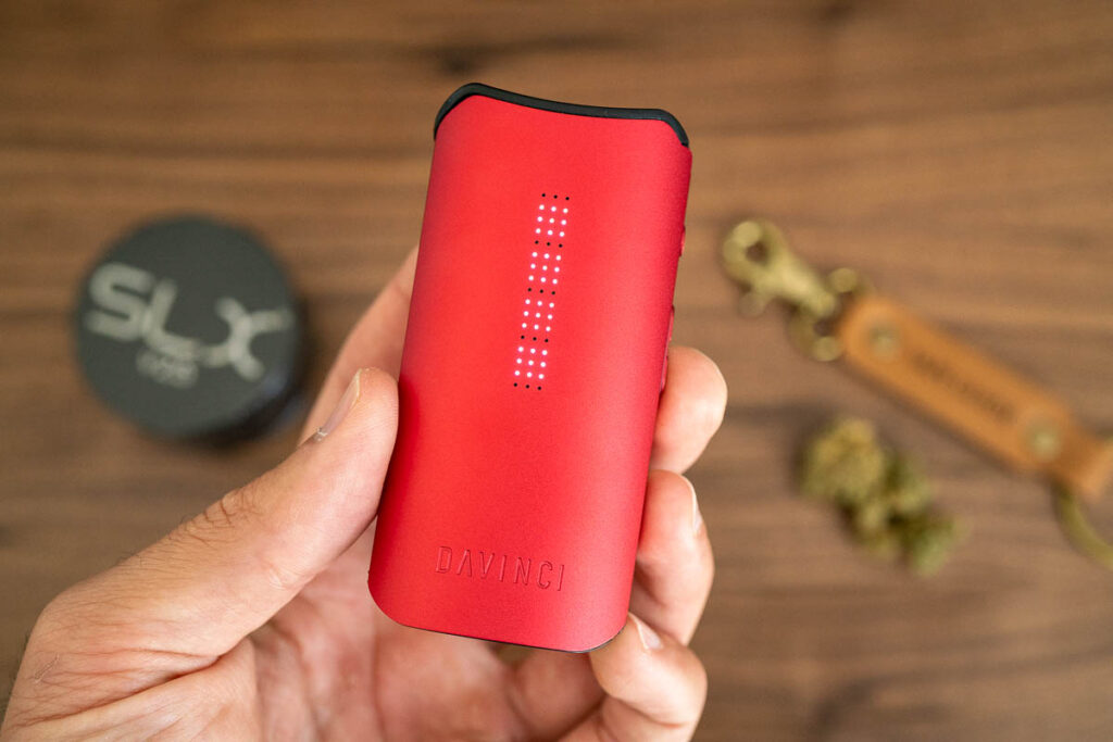 Close-up of a red Davinci vaporizer held in a hand, with accessories and herbs in the background