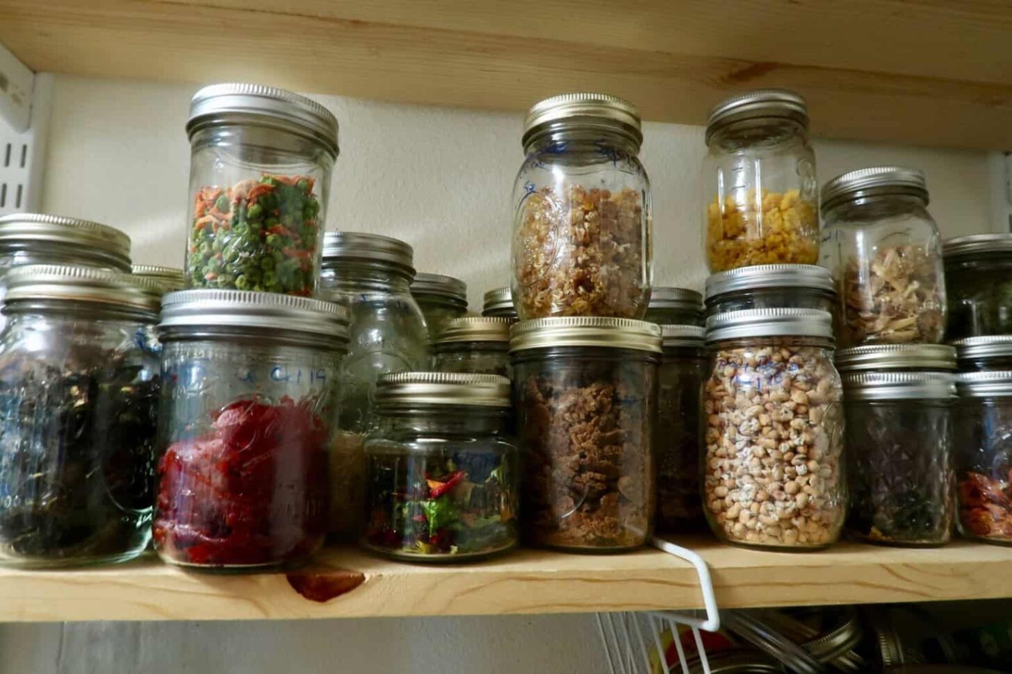 An assortment of glass jars filled with various dried foods, such as dried vegetables, herbs, and spices, organized on a wooden shelf.