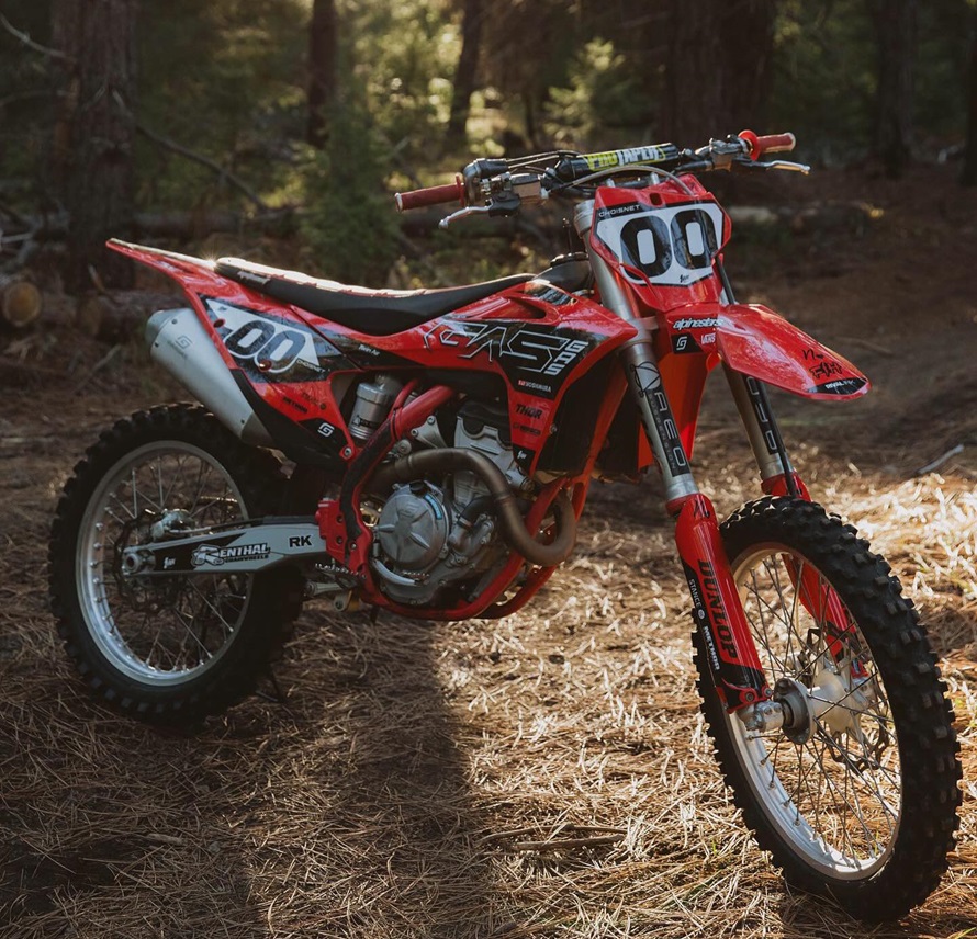  A red dirt bike with bold "00" race numbers on the front and side, parked in a wooded area. The bike features a custom MX graphics kit with a dynamic design, highlighting its rugged build and ready-for-adventure look.