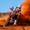 A motocross rider in full gear rides aggressively through a sand dune, creating an impressive dust trail behind. The red dirt bike stands out with its customized MX graphics kit, displaying the number "303" on the front plate, enhancing the bike’s sleek and competitive aesthetic.