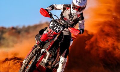 A motocross rider in full gear rides aggressively through a sand dune, creating an impressive dust trail behind. The red dirt bike stands out with its customized MX graphics kit, displaying the number "303" on the front plate, enhancing the bike’s sleek and competitive aesthetic.