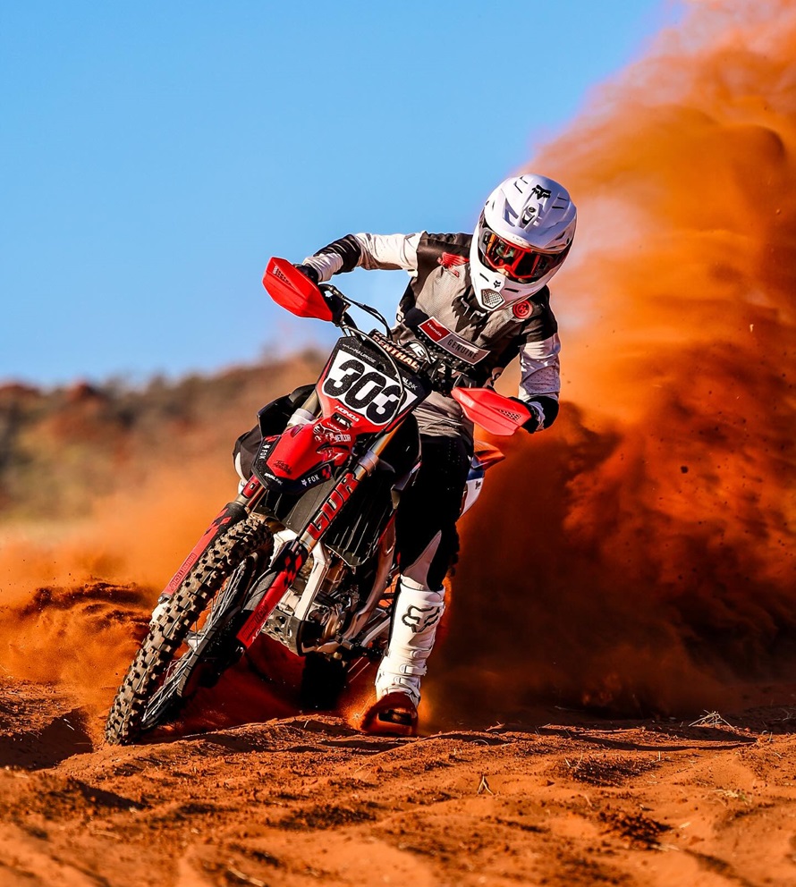 A motocross rider in full gear rides aggressively through a sand dune, creating an impressive dust trail behind. The red dirt bike stands out with its customized MX graphics kit, displaying the number "303" on the front plate, enhancing the bike’s sleek and competitive aesthetic.